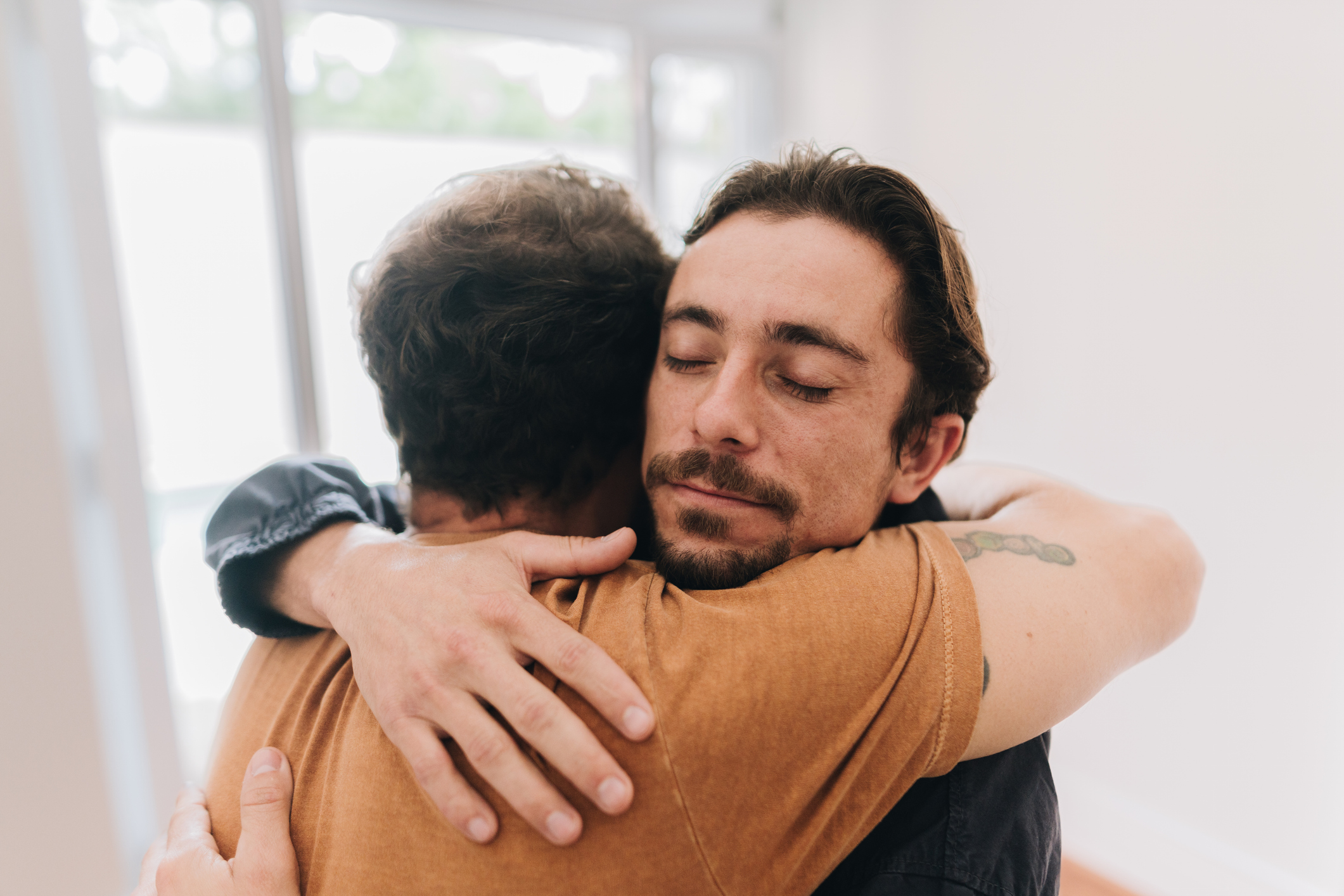 Two men hugging tightly, one with eyes closed, expressing care, empathy, and emotional healing.