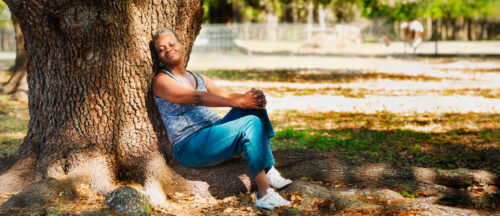 Older woman sitting peacefully against a tree in a sunlit park, appearing calm and reflective—suggesting a moment of self-acceptance or recovery.