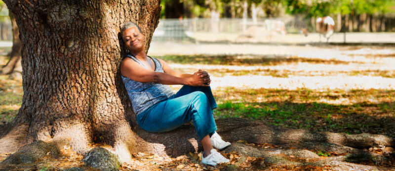 Older woman sitting peacefully against a tree in a sunlit park, appearing calm and reflective—suggesting a moment of self-acceptance or recovery.