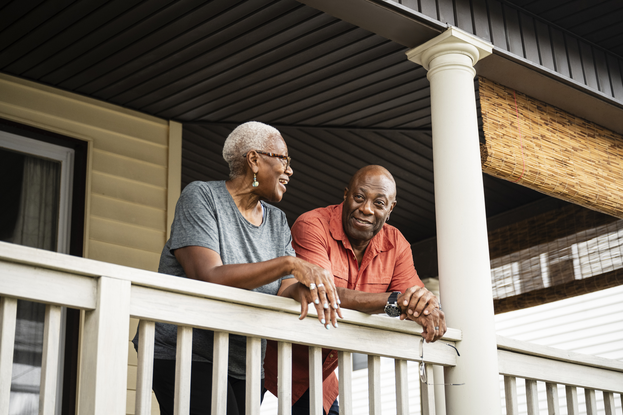 Older couple leaning on a porch railing, smiling and talking warmly, representing connection, support, and emotional well-being after experiencing verbal or emotional challenges.