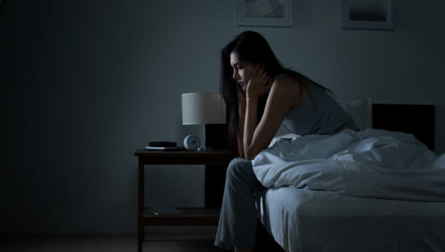 Woman sitting on the edge of a bed in a dark room with her head resting on her hand, conveying sadness, loneliness, or emotional exhaustion—symbolizing the overlap between autism and depression.