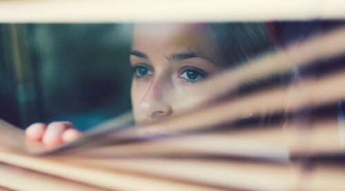 Close-up of a woman cautiously peering through window blinds, conveying anxiety, hesitation, or fear of being seen or judged.