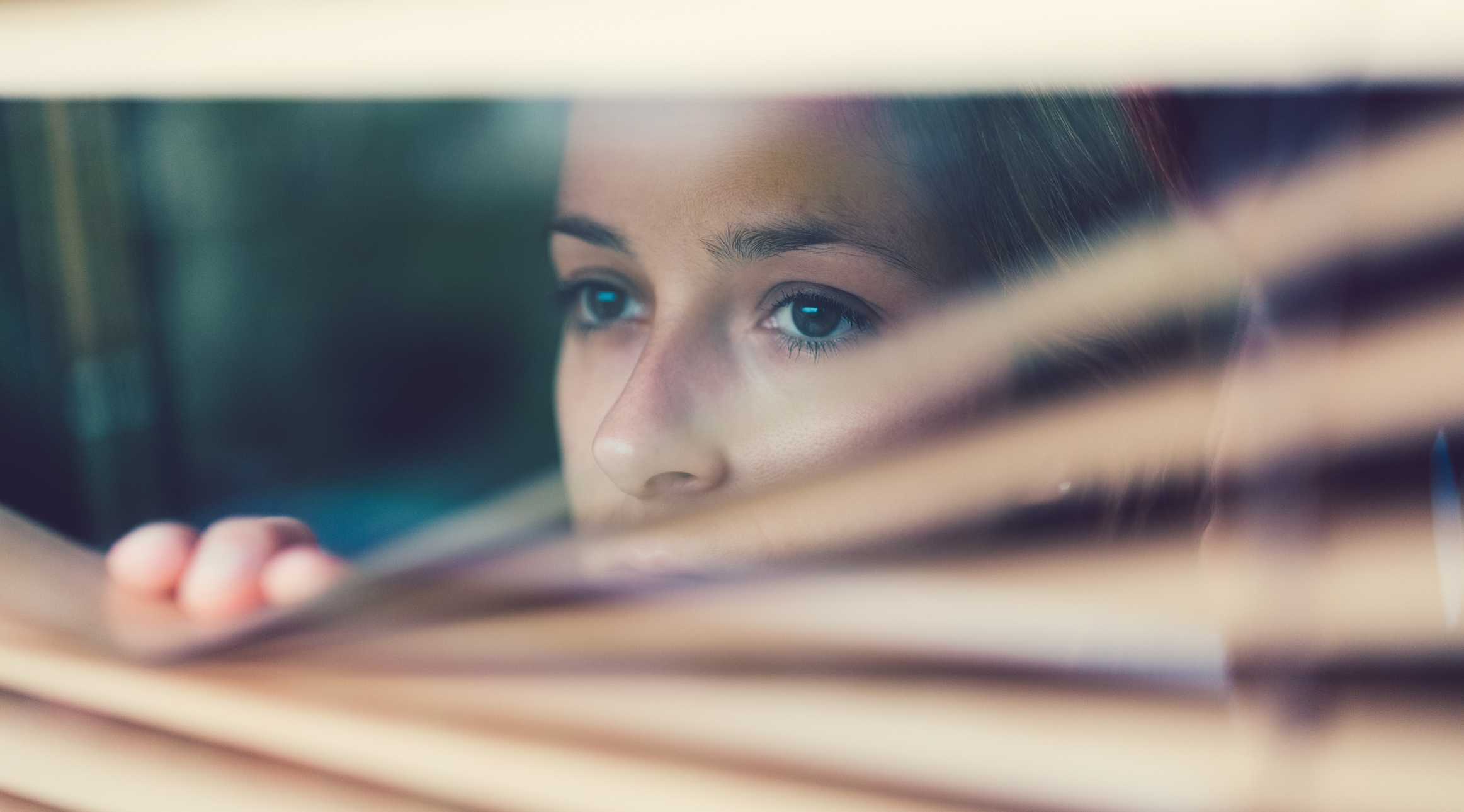 Close-up of a woman cautiously peering through window blinds, conveying anxiety, hesitation, or fear of being seen or judged.