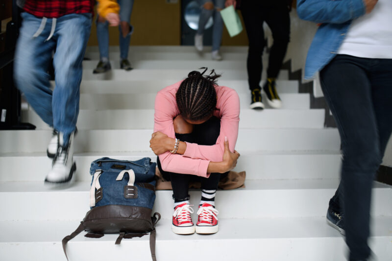 Stressed student sitting on school stairs with head down and arms wrapped around their knees, surrounded by other students walking past—symbolizing feelings of overwhelm and isolation at school.