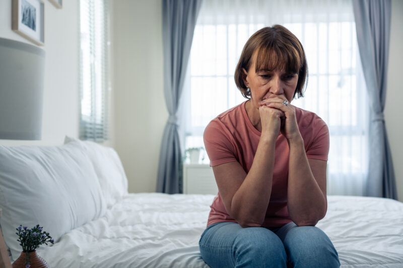 Middle-aged woman sitting on the edge of a bed, deep in thought, reflecting emotional distress linked to an inferiority complex.