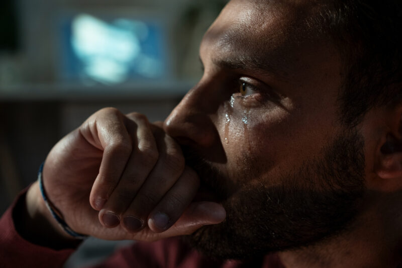 Close-up of a man crying in a dimly lit room, capturing the deep emotional pain and personal journey of grieving.