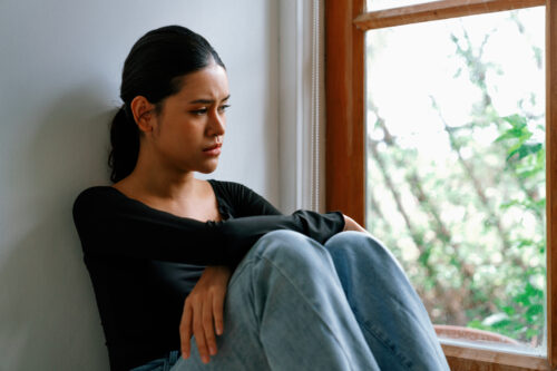 Young woman sitting alone by a window with a distressed expression, representing the emotional turmoil and isolation that can accompany PTSD and psychosis.