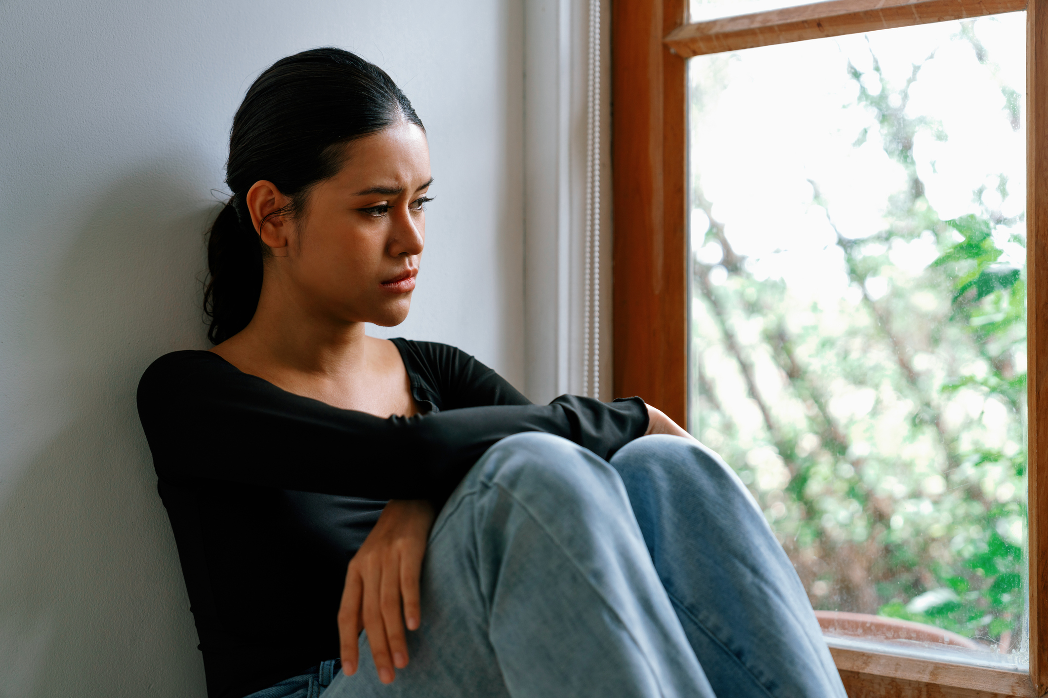 Young woman sitting alone by a window with a distressed expression, representing the emotional turmoil and isolation that can accompany PTSD and psychosis.