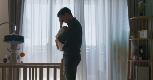 Stressed father holding a newborn in a dimly lit nursery, conveying emotional exhaustion and signs of depression.