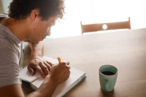 Man writing in a notebook at a table with a coffee mug, symbolizing reflection and personal exploration during an identity crisis.