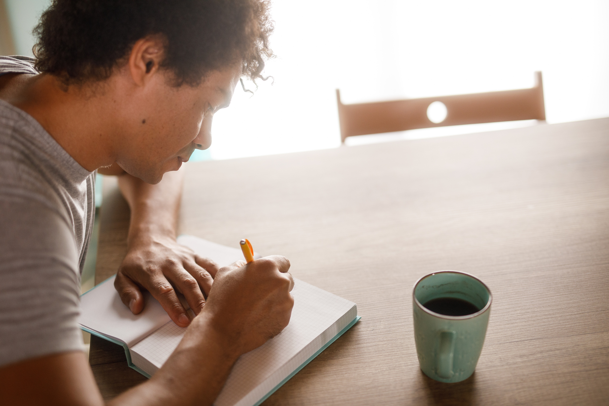 Man writing in a notebook at a table with a coffee mug, symbolizing reflection and personal exploration during an identity crisis.