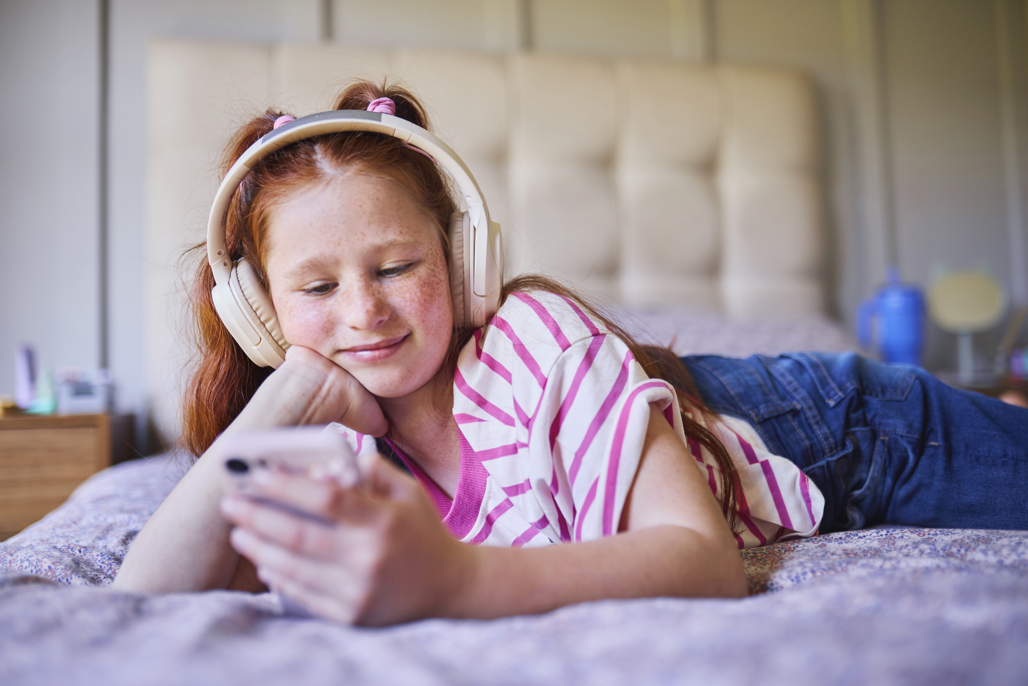 Young girl with red hair lying on her bed, wearing headphones and smiling at her phone, representing a relaxed moment of emotional well-being supported by personalized, holistic care.