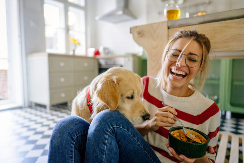 Smiling woman sitting on the kitchen floor with a warm meal and her dog by her side—capturing a cozy, grounding moment of self-care and emotional relief, helpful in managing PMDD.