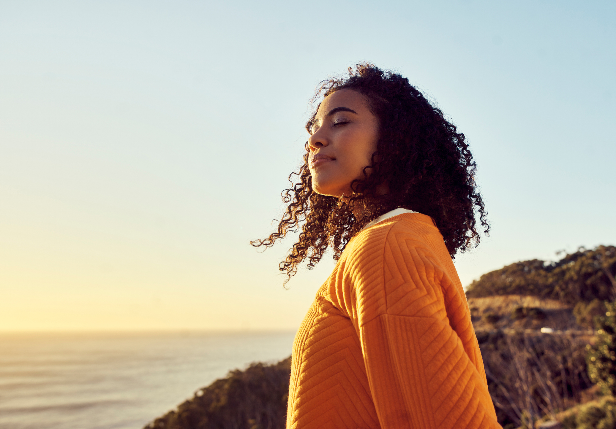 Young woman standing peacefully near the ocean at sunset with eyes closed, taking a deep breath and soaking in the calm—symbolizing healing, mindfulness, and emotional clarity.