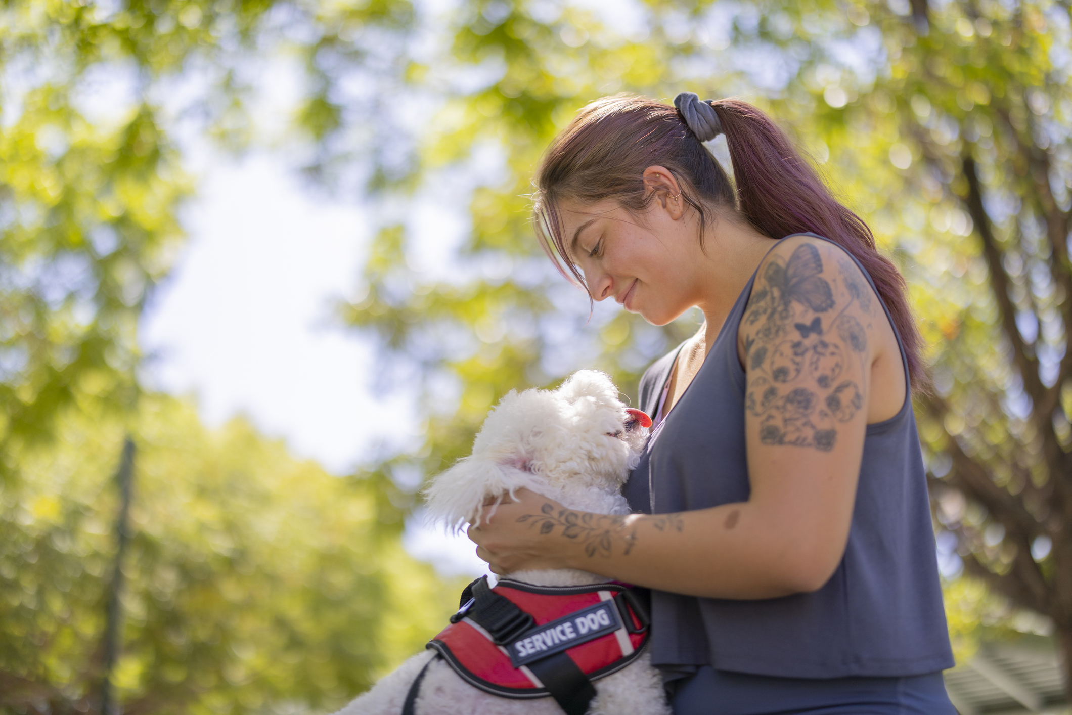 A woman with tattoos smiles while holding her small white service dog outdoors.