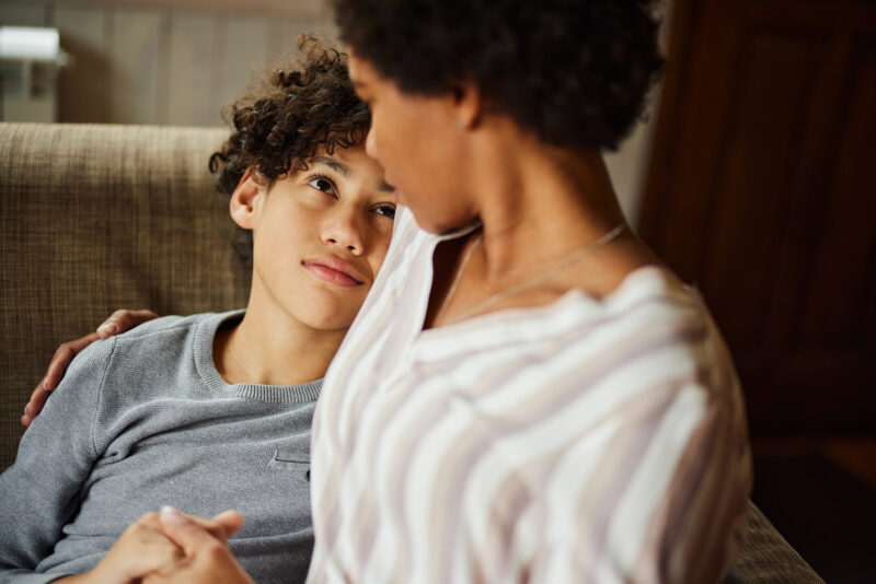 Teen sitting on a couch, gazing up at a supportive adult who holds their hand, symbolizing trust, care, and emotional connection during a vulnerable moment.