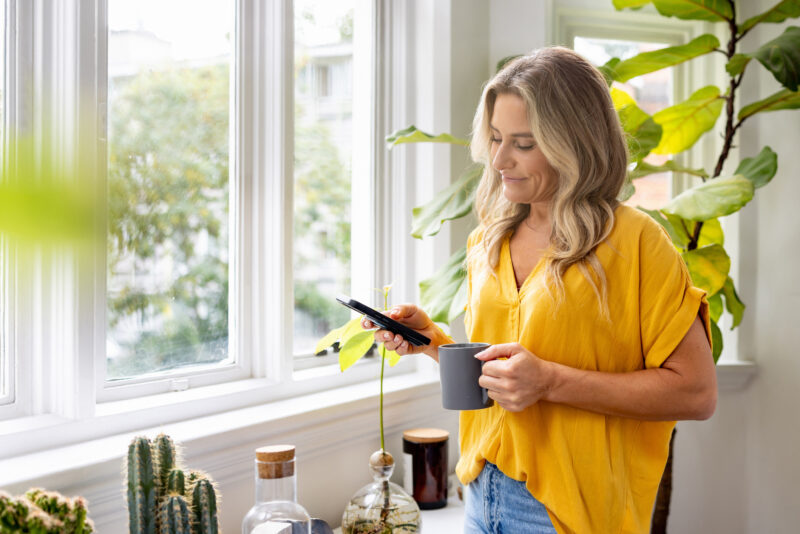 Woman standing by a sunny window, holding a mug and checking her phone with a calm expression—capturing a quiet moment of self-reflection, a reminder to pause and consider taking a mental health day.