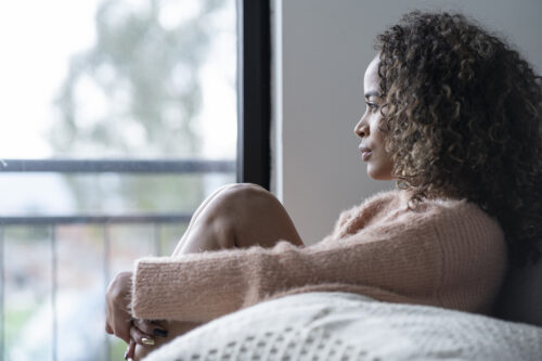 Woman sitting by a window with her knees pulled close, gazing outside with a thoughtful expression, reflecting introspection or emotional processing—key aspects of behavioral health.