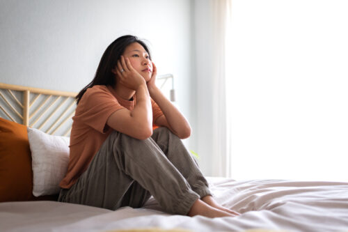 Young woman sitting indoors with her face in her hands, staring ahead pensively, suggesting inner conflict or body image concerns.