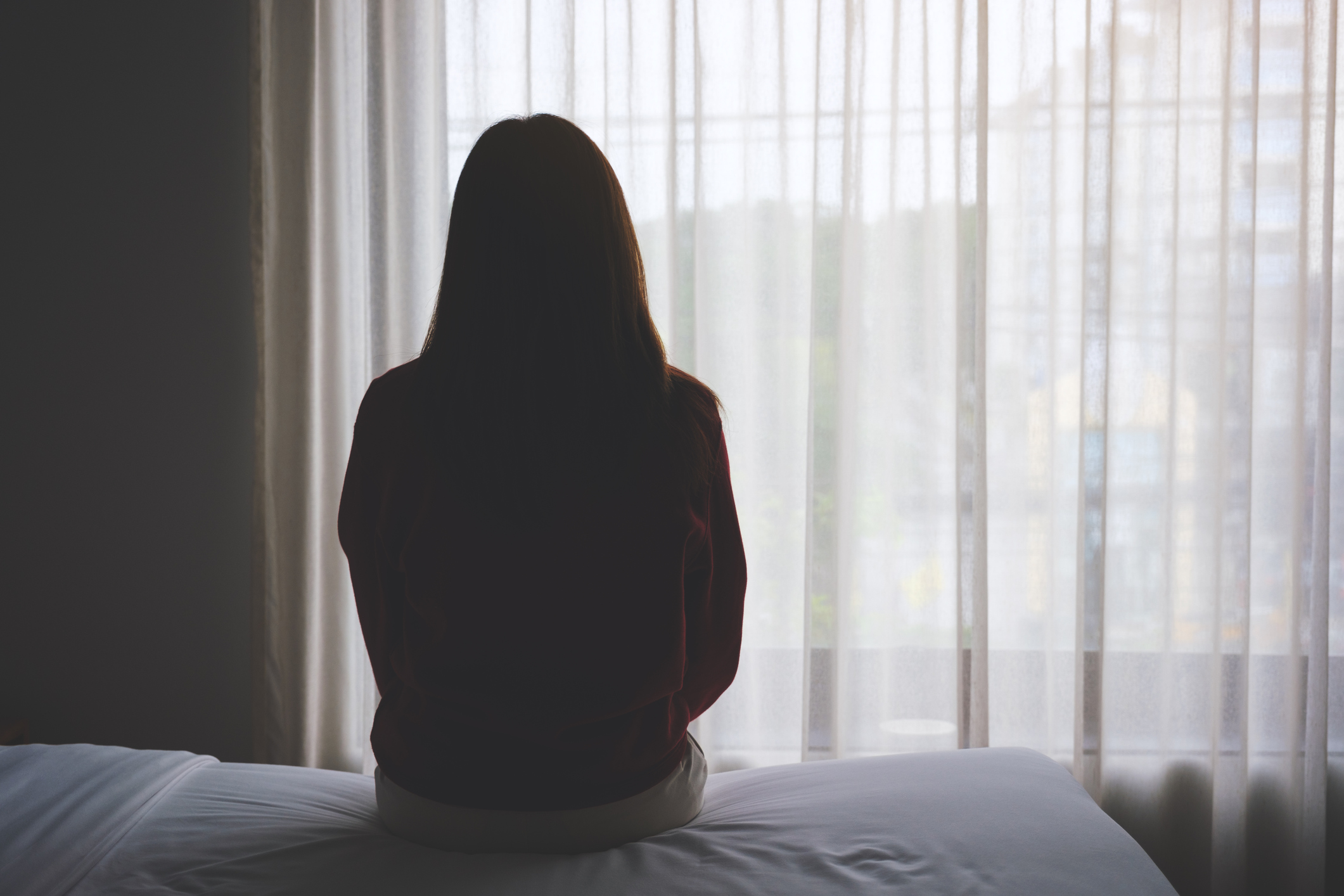 Silhouette of a person standing alone and looking out a window with sheer curtains, evoking a quiet, persistent sense of emotional heaviness often associated with dysthymia or chronic depression.