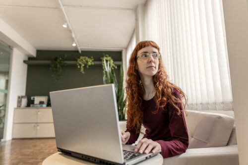 Person with long red hair sitting at a laptop, looking off to the side with a serious expression, suggesting mental fatigue or sensory overload.