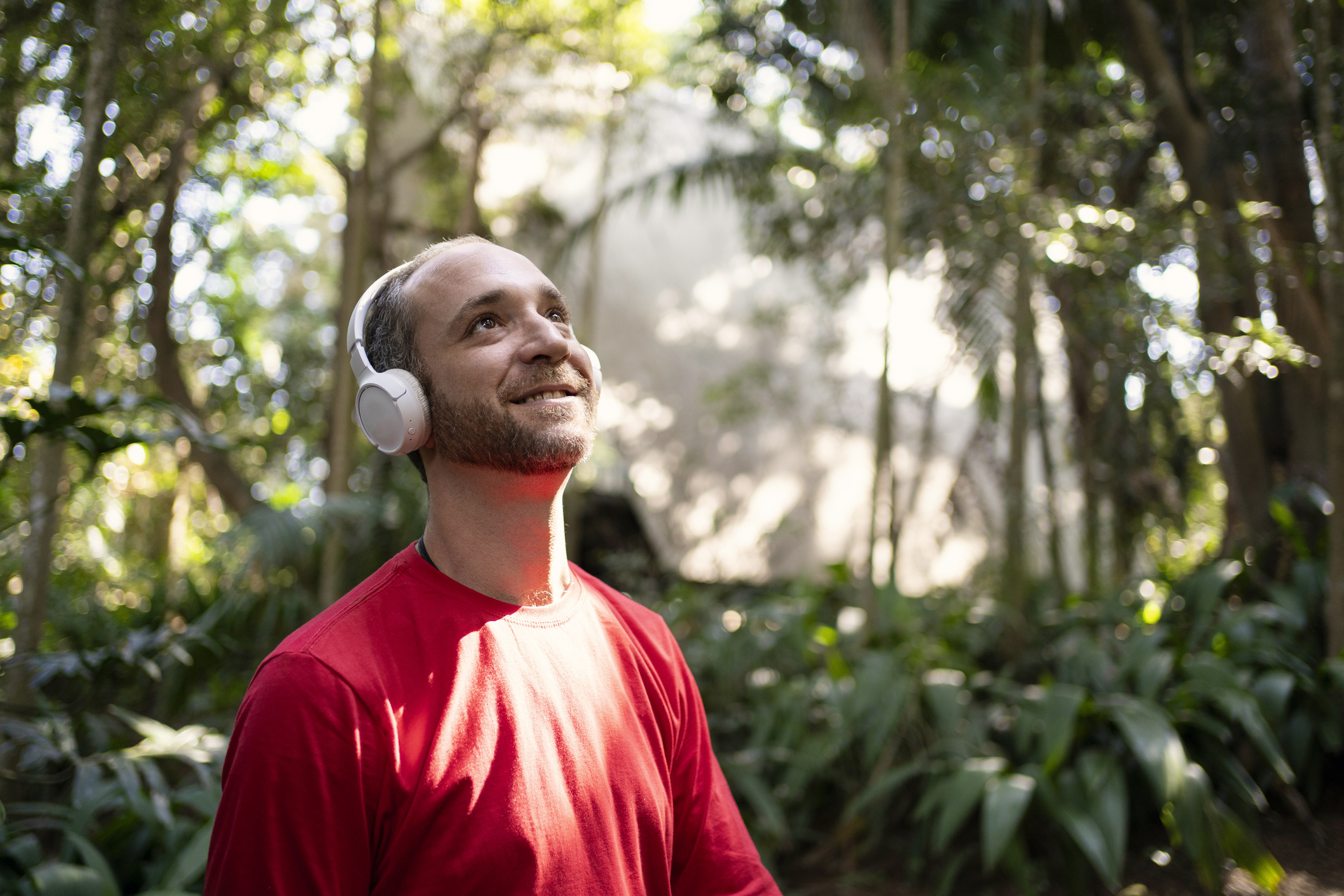 Smiling man wearing headphones and a red shirt, standing in nature and looking up, symbolizing moments of peace or sensory relief after burnout.