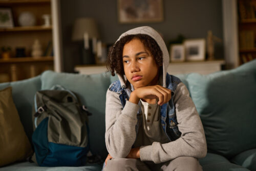 Teenager sitting on a couch with a withdrawn expression, wearing a hooded sweatshirt, suggesting emotional discomfort or early signs of depression.