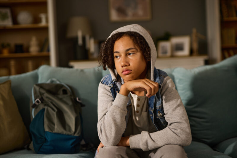 Teenager sitting on a couch with a withdrawn expression, wearing a hooded sweatshirt, suggesting emotional discomfort or early signs of depression.
