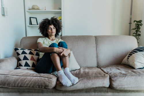 Woman sitting on a couch with knees pulled to her chest, staring off in thought with a subdued expression, representing the quiet, persistent weight of dysthymia.