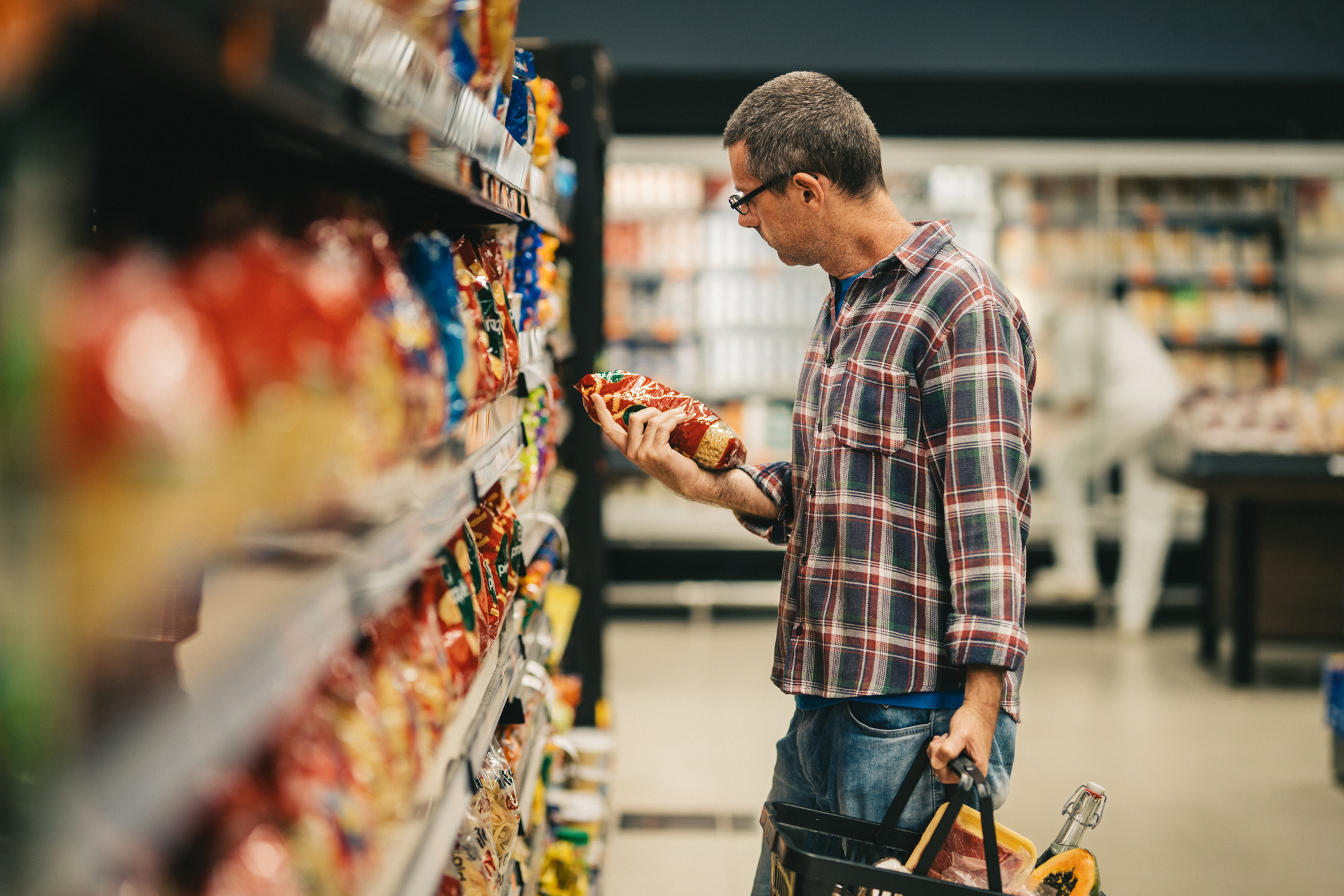 Man wearing a plaid shirt closely examining a bag of chips in a grocery store aisle, symbolizing mindful or conflicted food choices.