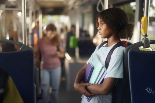 Young girl sitting alone with a serious expression, holding school supplies tightly to her chest, possibly overwhelmed in a noisy or overstimulating environment—suggestive of sensory sensitivity.