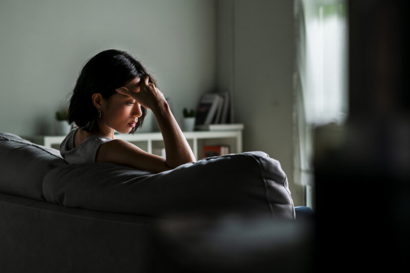 Young woman sitting on a couch with her hand on her forehead, eyes closed in visible tension, capturing the physical and emotional weight of stress or anxiety.