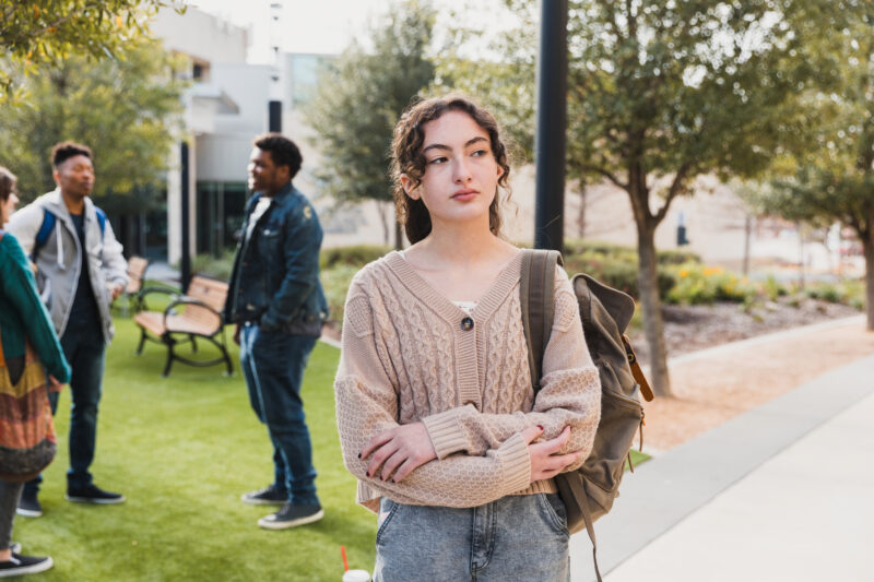 Teen girl standing alone on campus looking withdrawn while others socialize nearby, reflecting increased risks of substance use, isolation, and peer pressure during summer months.