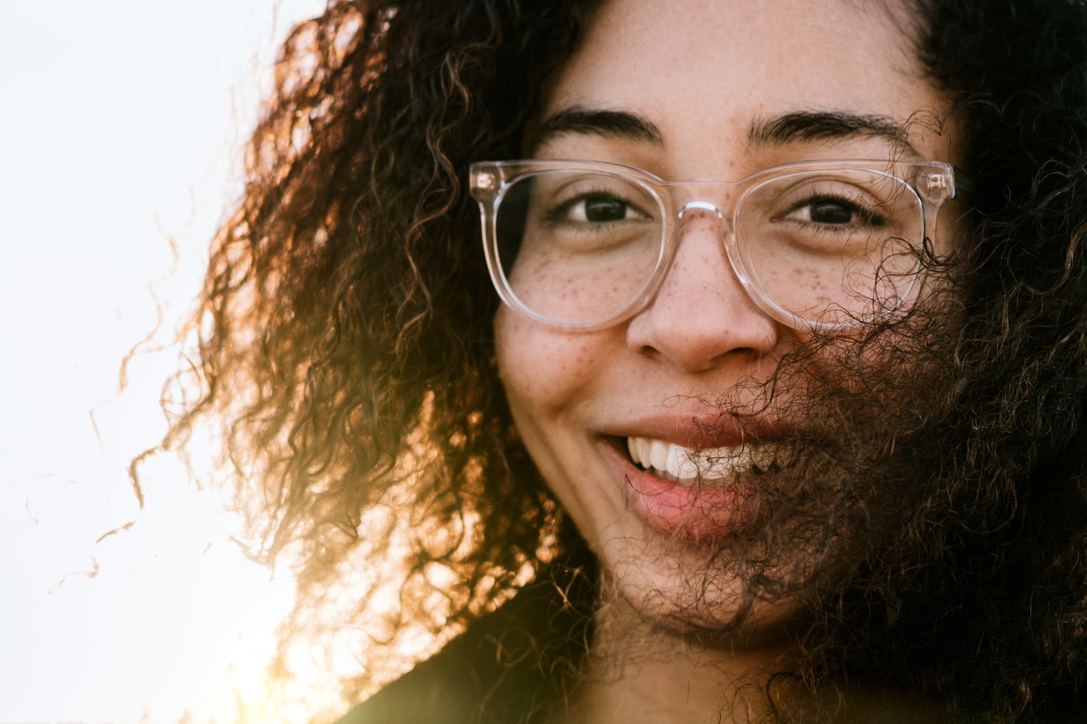 A naturally beautiful mixed race woman smiles for a portrait, the sun shining from behind.  Her clear glasses frames highlight her freckles.