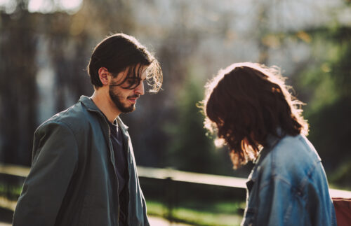 Two people standing outside during a serious conversation, both looking down with visible emotional tension. The image captures the emotional weight and disconnection that can arise from self-sabotage in relationships.