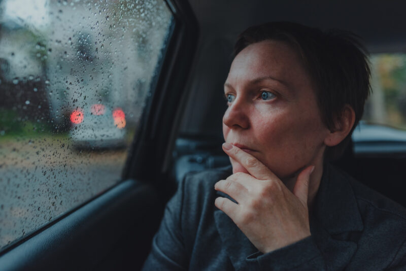 Person sitting in a car, staring out a rain-covered window with a distant, pensive expression and hand near their mouth. The image captures a quiet moment of emotional intensity, evoking the feeling of being mentally pulled back into a past experience or traumatic memory.