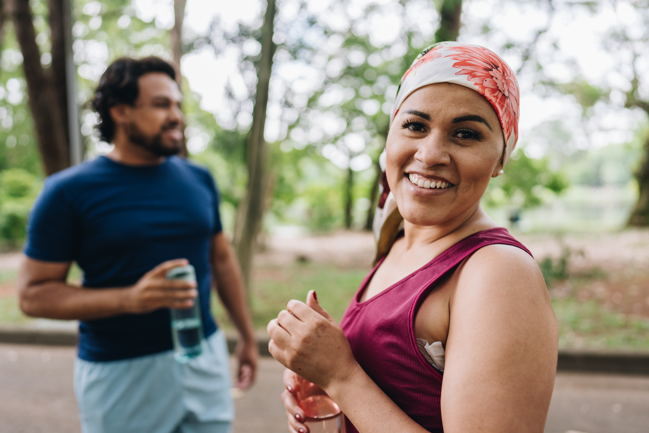 Smiling woman wearing a headscarf and athletic wear holding a water bottle during a walk in the park, with a man in the background. The image reflects connection, recovery, and healing in a supportive environment.