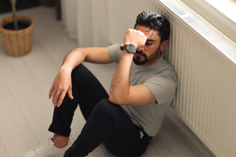 Man sitting on the floor with eyes closed and arm resting over his face, appearing emotionally overwhelmed. The image reflects deep psychological distress, capturing the isolation and disorientation that can accompany trauma-induced psychosis.