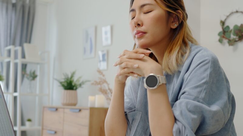 Person sitting with eyes closed and hands clasped, appearing deep in thought or emotionally overwhelmed. The image conveys a moment of inner reflection or emotional processing, often linked to trauma-related behaviors like oversharing.