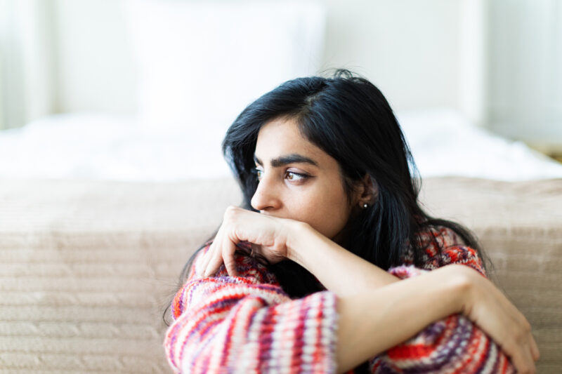 Woman sitting on a couch with her arms crossed over her knees, gazing off to the side with a distant, anxious expression. The image conveys emotional distress and deep rumination, reflecting the overlap between obsessive-compulsive disorder and more severe cognitive or perceptual disruptions like psychosis.