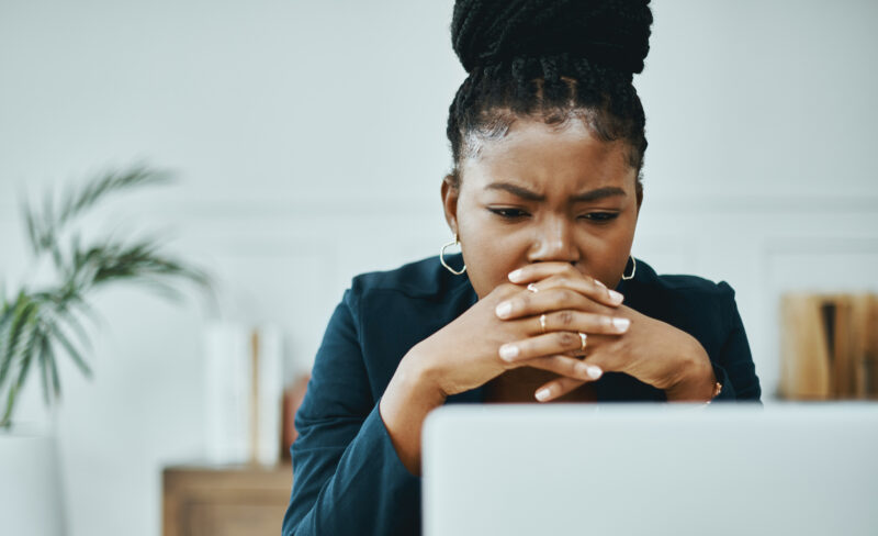 A woman sits with her hands clasped in front of her mouth, brows furrowed in deep concentration or emotional stress. She appears to be looking at a laptop screen, wearing a dark blazer and hoop earrings in a well-lit indoor setting.