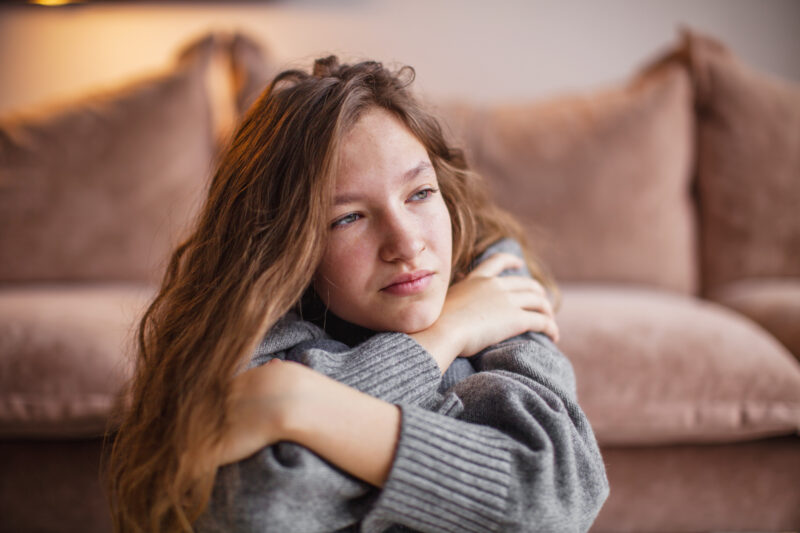 A teenage girl with long brown hair sits with her arms wrapped around her knees, gazing off to the side with a distant, pensive expression. She is wearing a cozy gray sweater, and the warm, soft-focus background suggests a home or comforting indoor environment.