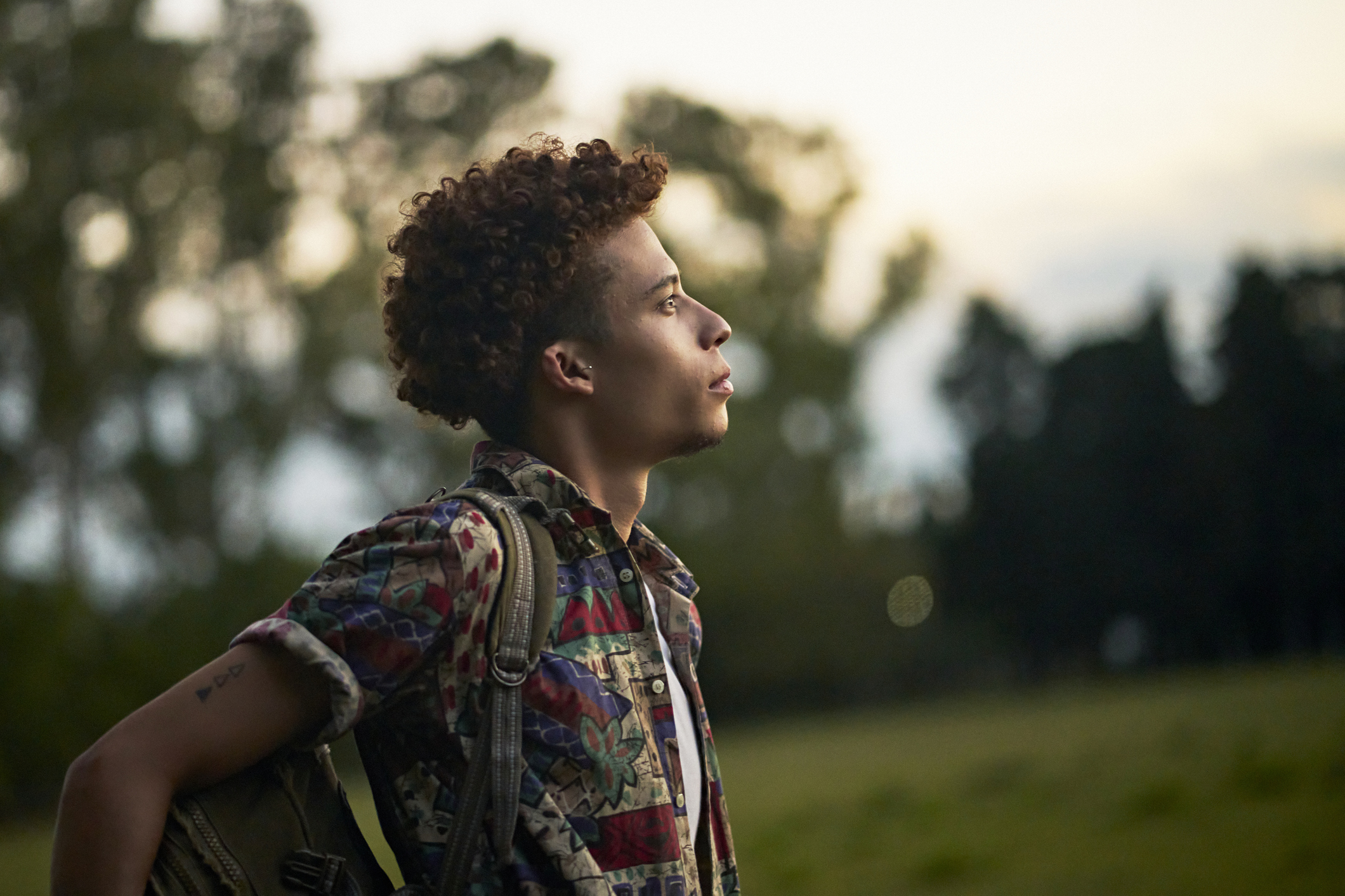 A young person with curly hair and a colorful patterned shirt stands in a grassy field at dusk, carrying a backpack and gazing upward with a thoughtful expression. The scene evokes themes of reflection, personal growth, and the search for stability—central to conversations around psychiatric medication for anxiety, depression, and ADHD.