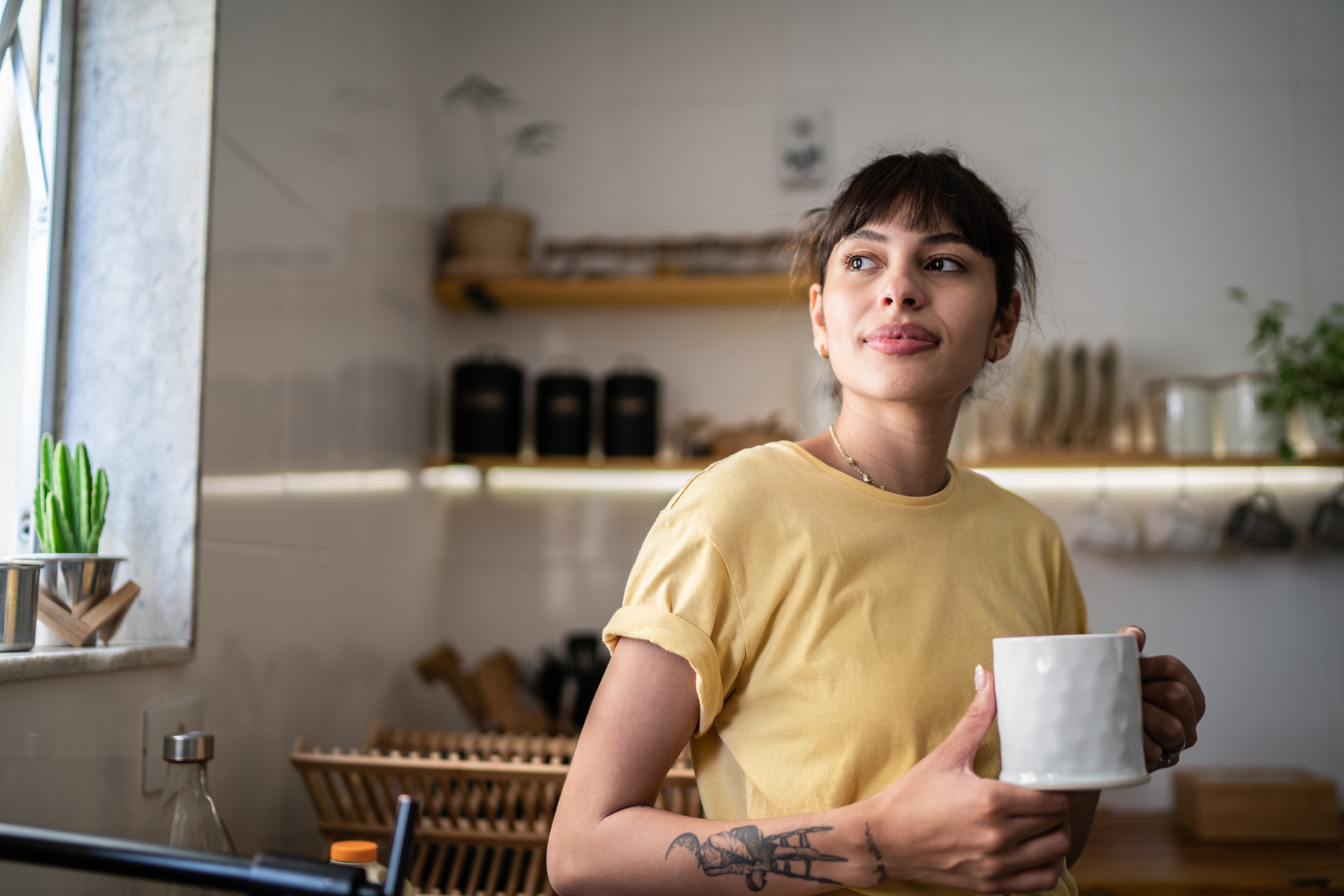 Young woman standing in a cozy kitchen holding a mug, with a calm, reflective expression. She appears grounded and at ease, illustrating the potential for anxiety relief when cortisol levels are balanced, according to therapists.
