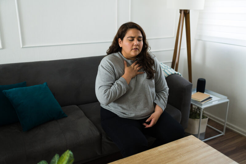 A woman sits on a couch indoors with her eyes closed and one hand on her chest, appearing to experience discomfort or distress. She is wearing a gray long-sleeve shirt, and the setting suggests a home environment with a white wall and floor lamp in the background.
