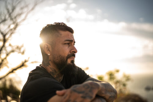 A man with tattoos and a beard sits outdoors at sunset, arms crossed and eyes looking into the distance with a serious, contemplative expression. His posture and expression reflect the emotional weight of navigating mental health conditions like anxiety, depression, or ADHD—topics psychiatrists often address through medication.