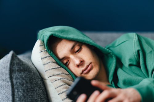 A young man in a green hoodie lies on a couch, staring at his phone with a tired or low-energy expression. His body language suggests emotional withdrawal, capturing the depressive symptoms that can occur after quitting smoking.