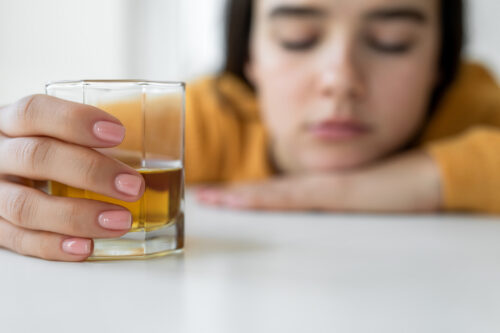 A close-up of a young woman resting her head on a table, eyes closed, with a tired or somber expression. Her hand is holding a glass of amber liquid, possibly alcohol. The focus is on the glass, while her face is blurred in the background.