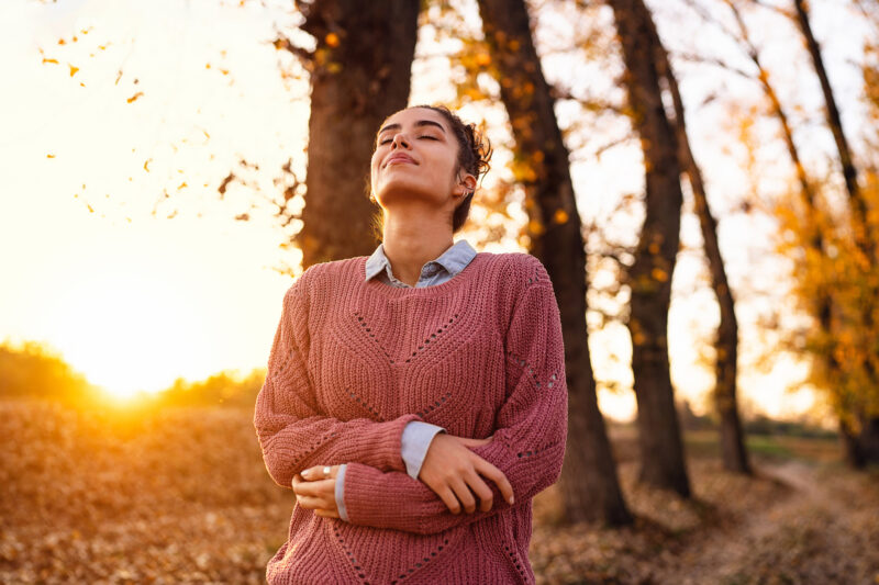 A young woman stands outdoors in a forest during autumn, eyes closed and arms gently crossed, wearing a pink knitted sweater over a collared shirt, basking in the warm golden sunlight.