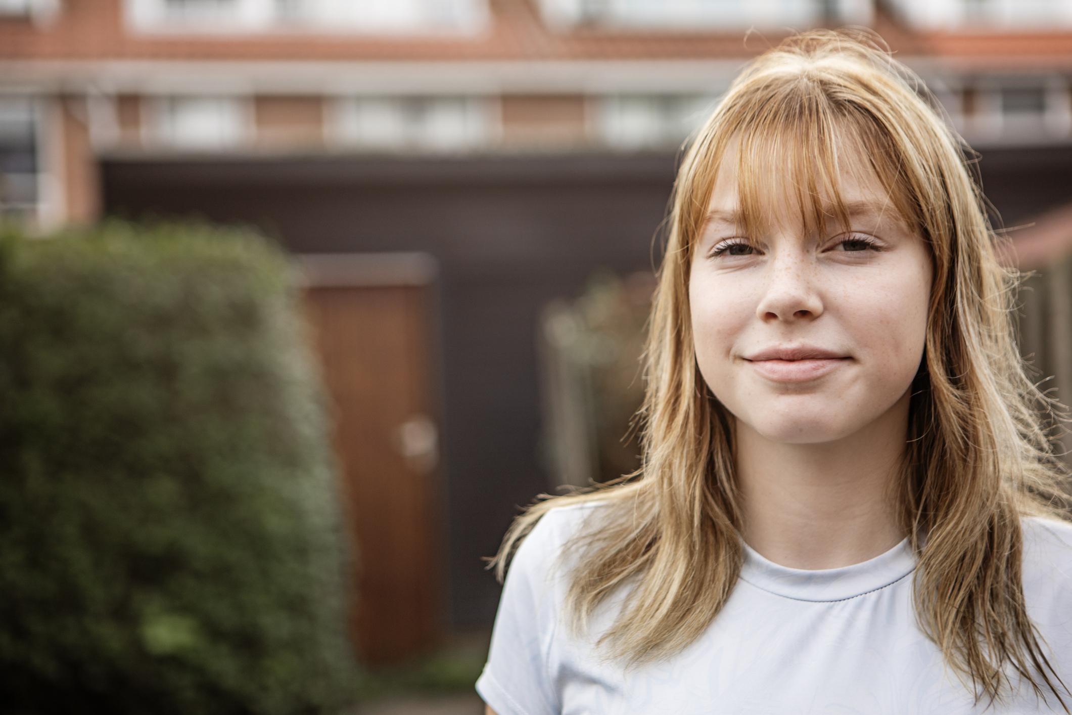 A teenage girl with light red hair and bangs stands outside, looking directly at the camera with a slight smile. She wears a pale blue shirt, and the blurred background shows a building with dark walls and a tiled roof.